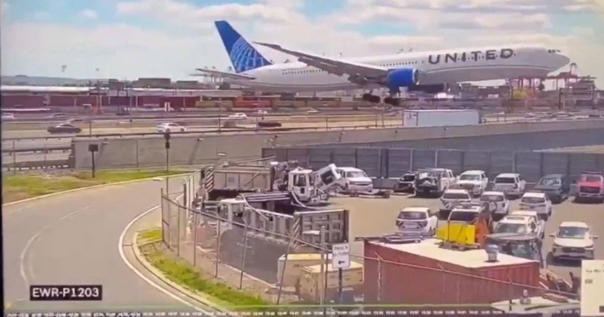 United Airlines plane landing at Newark Liberty International Airport with parked vehicles and a clear blue sky in the background.