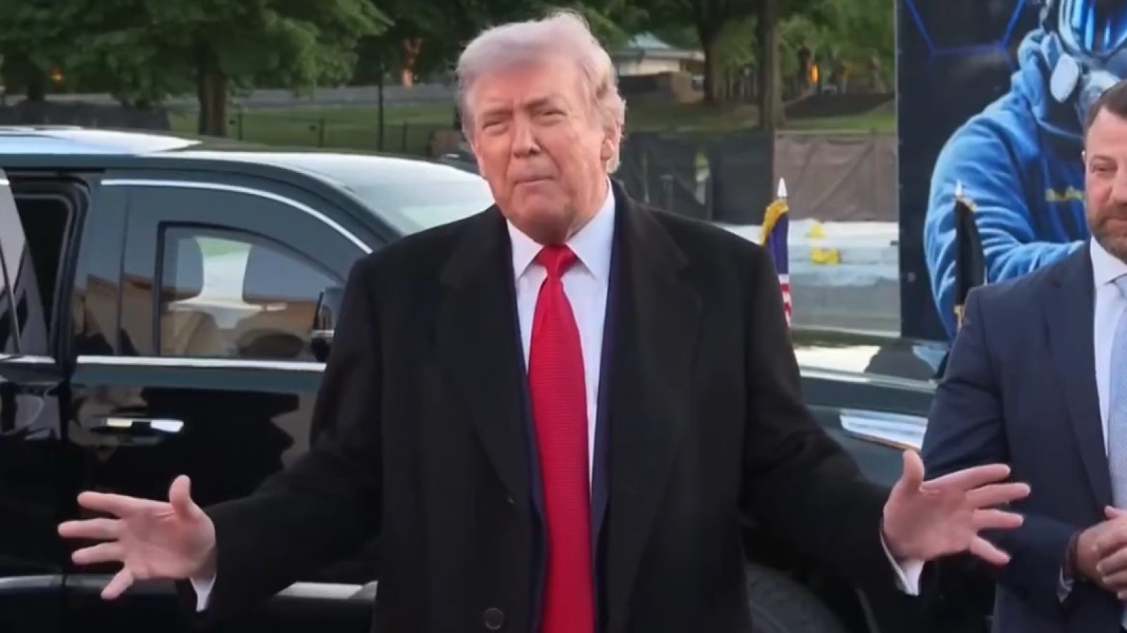Donald Trump gestures while speaking outside, with a black SUV in the background and a man in a suit beside him.