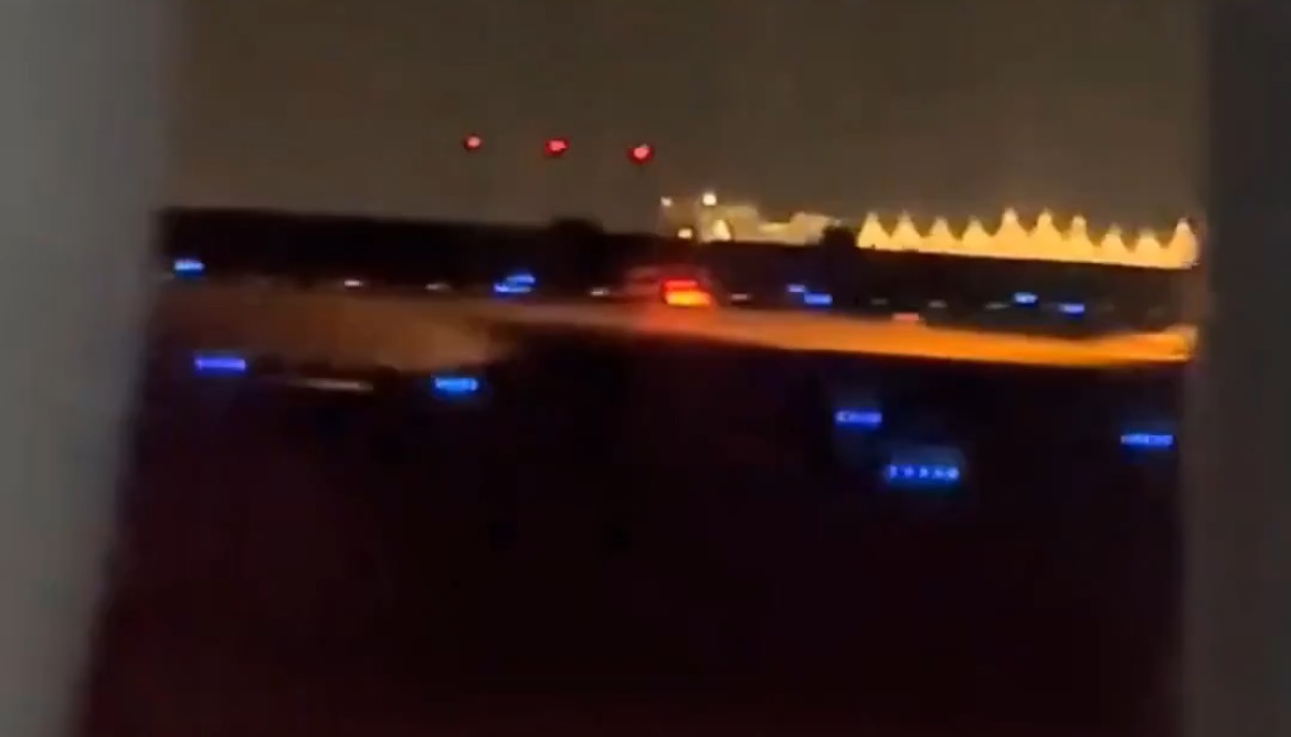 Nighttime airport runway view with illuminated taxiway lights and red signal lights in the background.