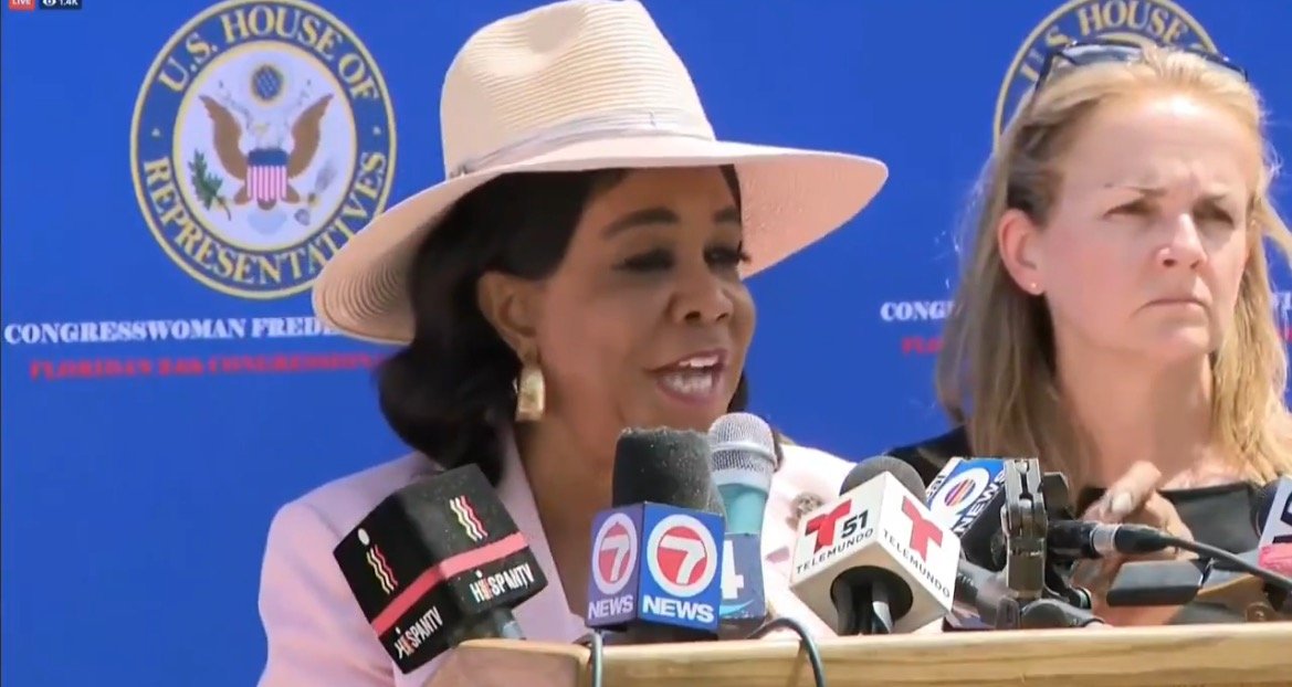 Congresswoman speaking at a press conference with various media microphones, highlighting important issues in front of a U.S. House of Representatives backdrop.