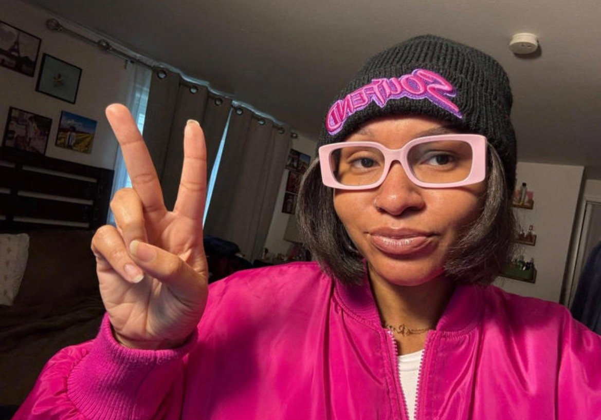 Person wearing a black beanie and pink glasses makes a peace sign while posing in a brightly colored jacket in a cozy indoor setting.