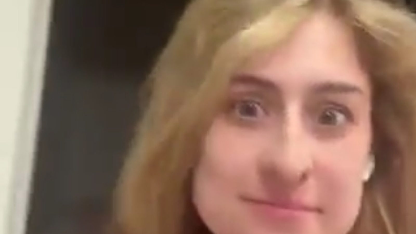 Young woman with blonde hair and earbuds smiling at the camera, capturing a candid moment in a home setting.