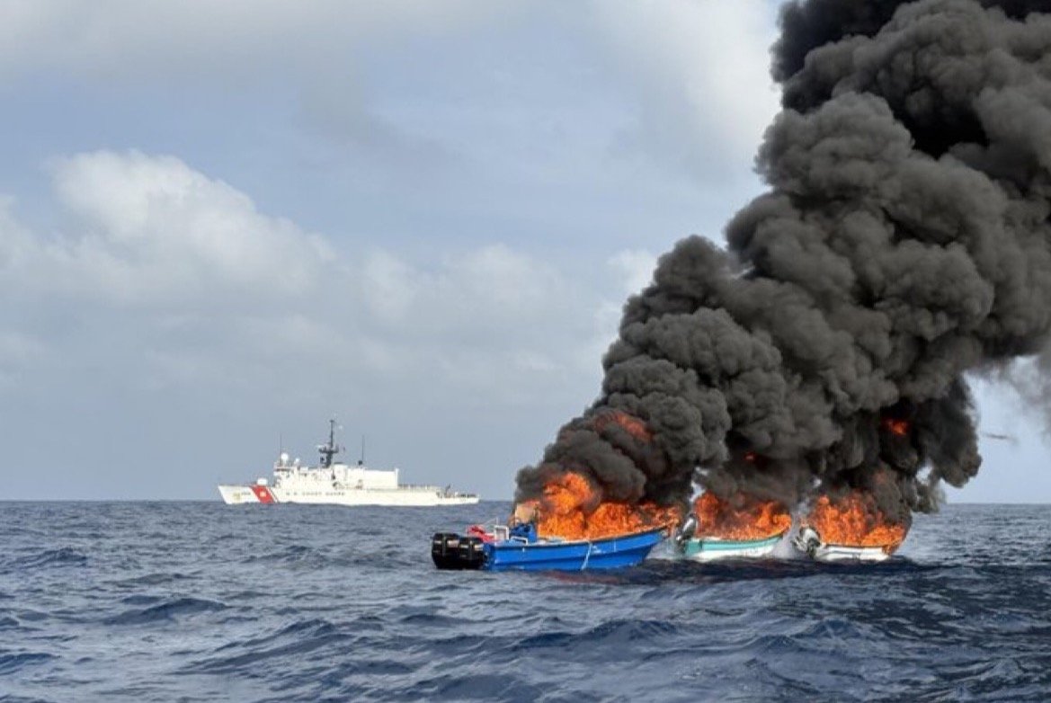 Burning fishing boats create thick black smoke at sea with a coast guard vessel in the background.