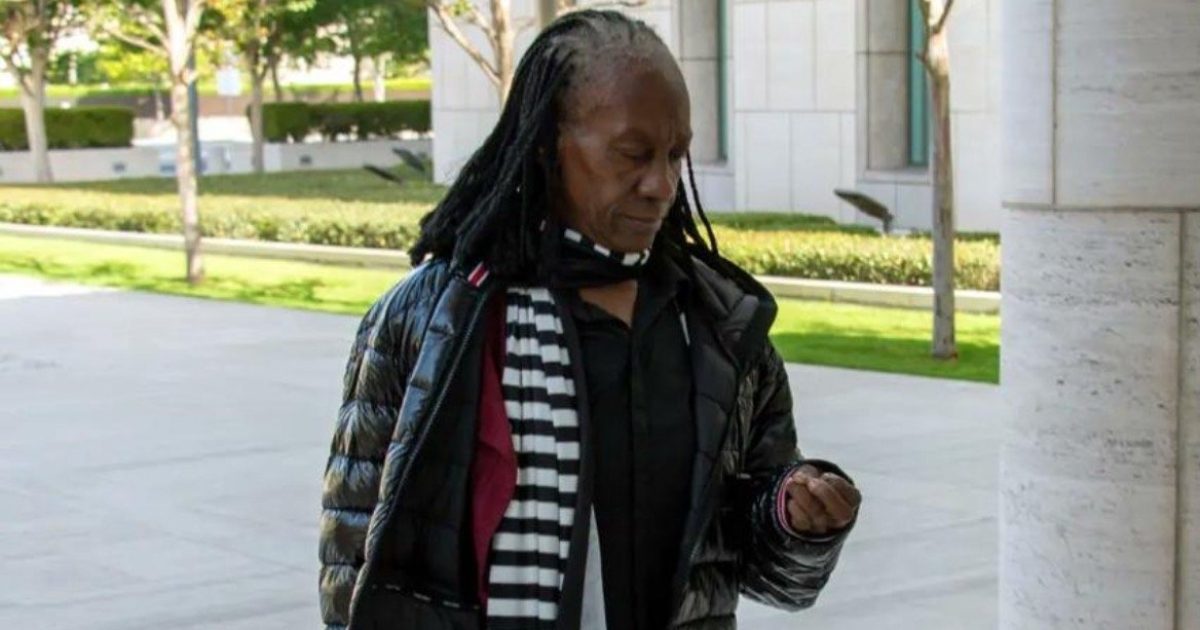 Person with long black hair and a striped scarf checks their watch while standing outdoors in a modern architectural setting.