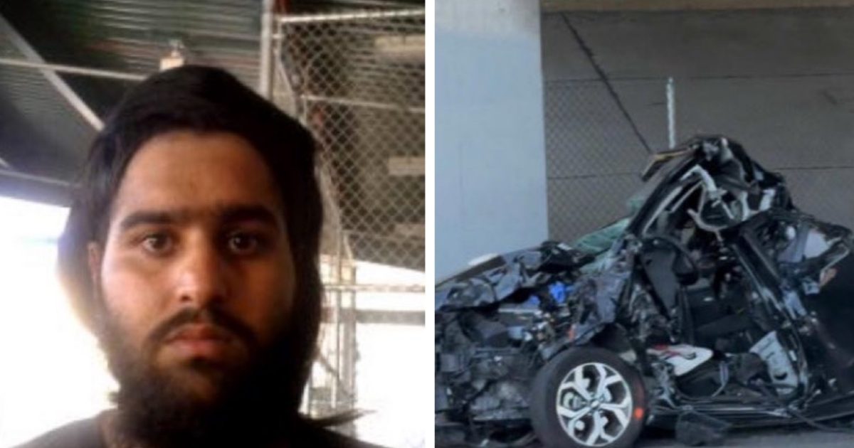 A man with a beard stands in front of a badly damaged car, highlighting the aftermath of a serious accident.