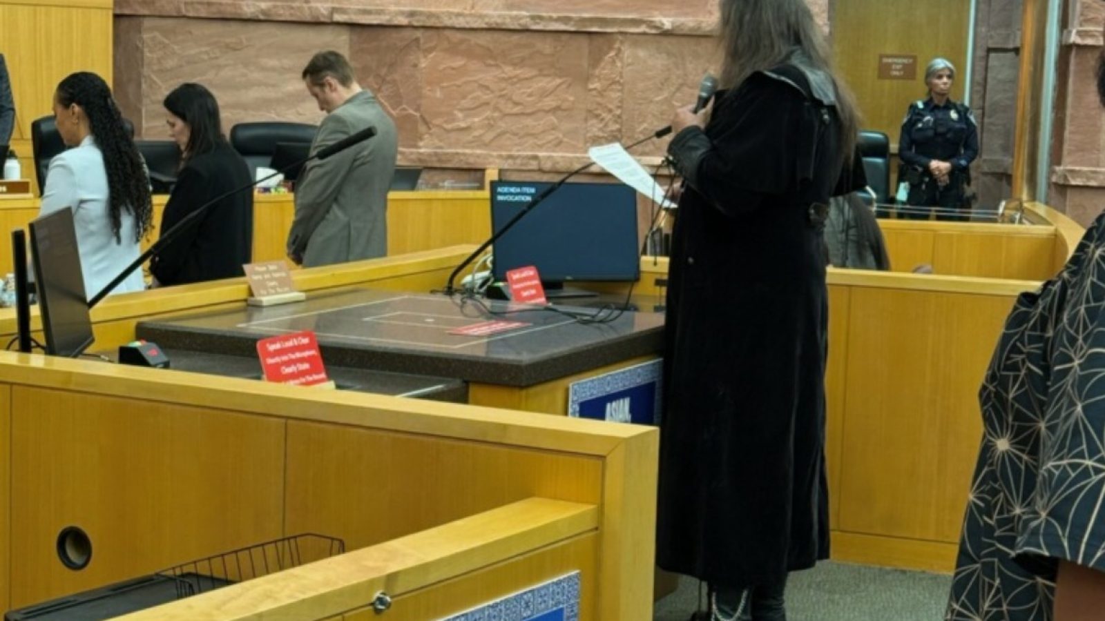 A person in a black outfit speaks at a public meeting in a courtroom, with attendees listening attentively and a police officer in the background.