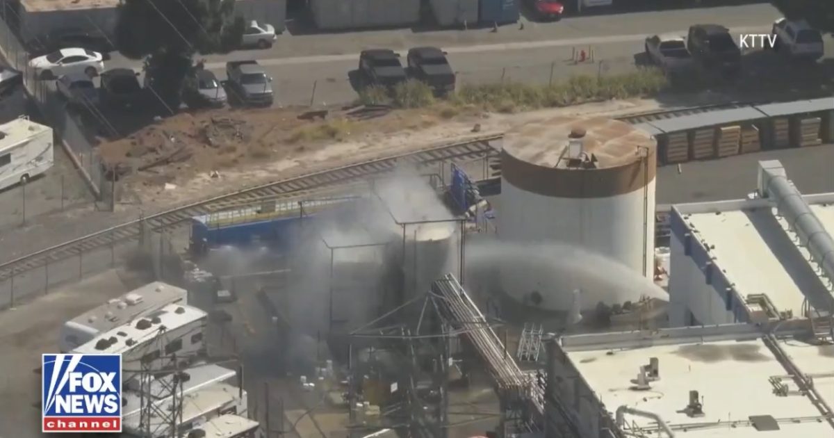 Industrial site with smoke rising from storage tanks, surrounded by parked vehicles and railway tracks in the background.