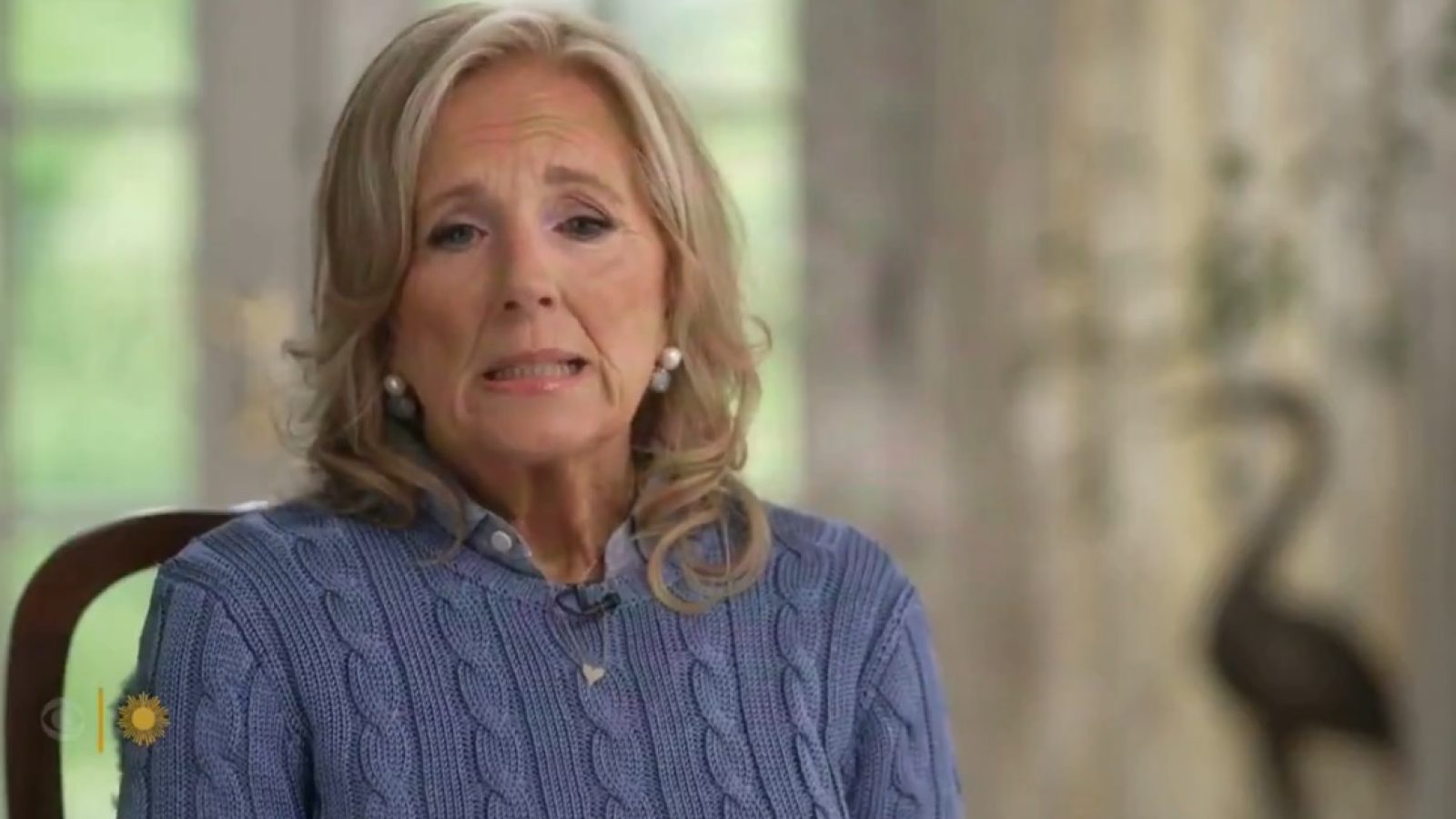 Woman with long blonde hair wearing a blue sweater, sitting indoors with a thoughtful expression, surrounded by natural light and soft decor.