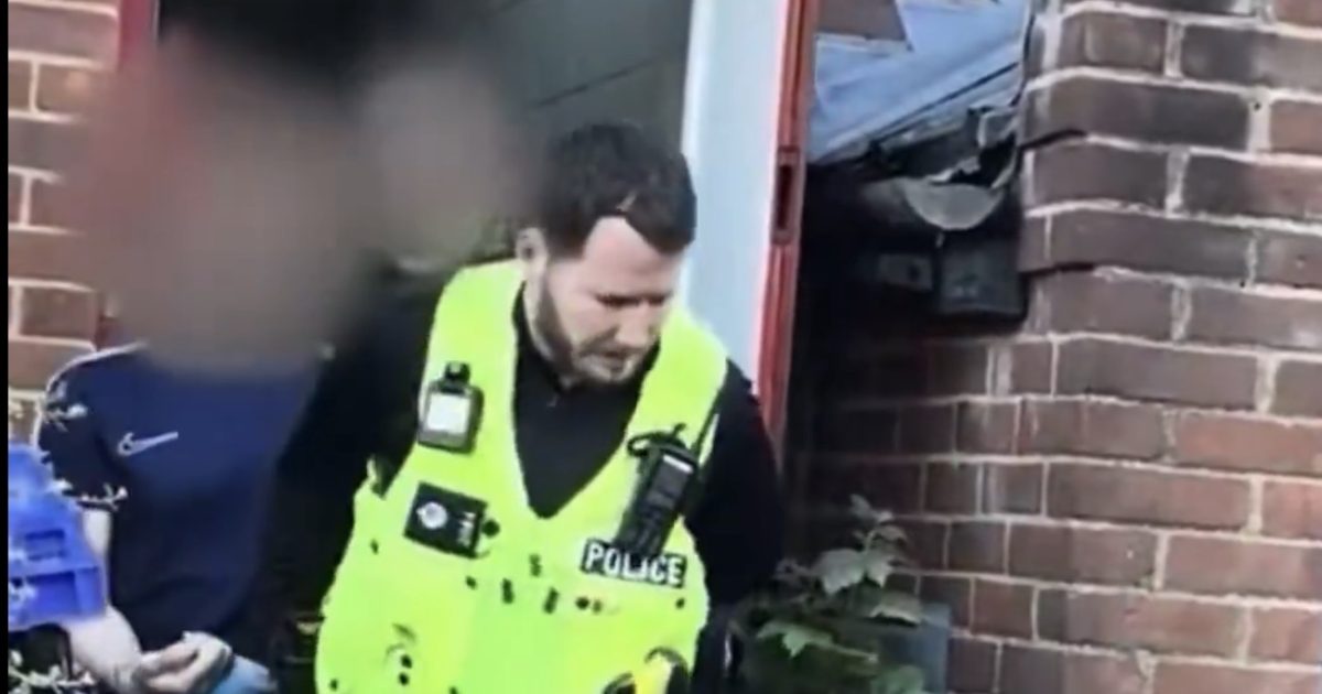Police officer in a high-visibility vest apprehending an individual near a residential entrance surrounded by brick walls and greenery.