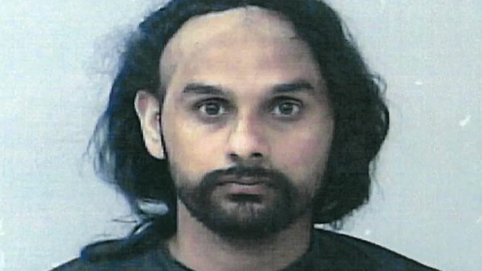 Mugshot of a male individual with long dark hair and a distinctive bald patch, wearing a black shirt against a plain background.