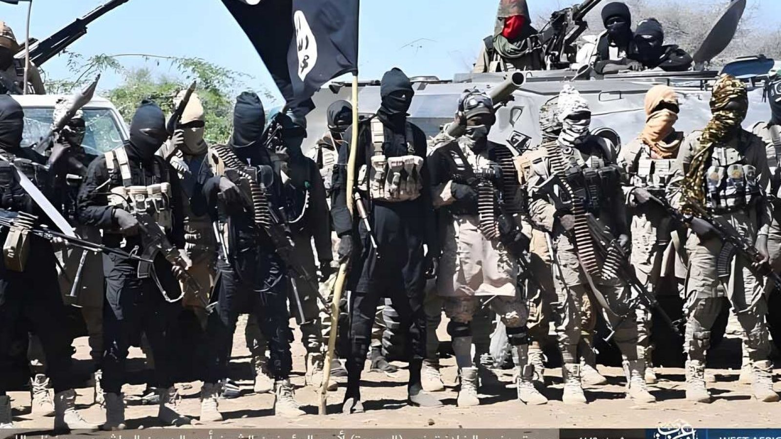 Militant group members in tactical gear displaying weapons and flags in a desert environment.