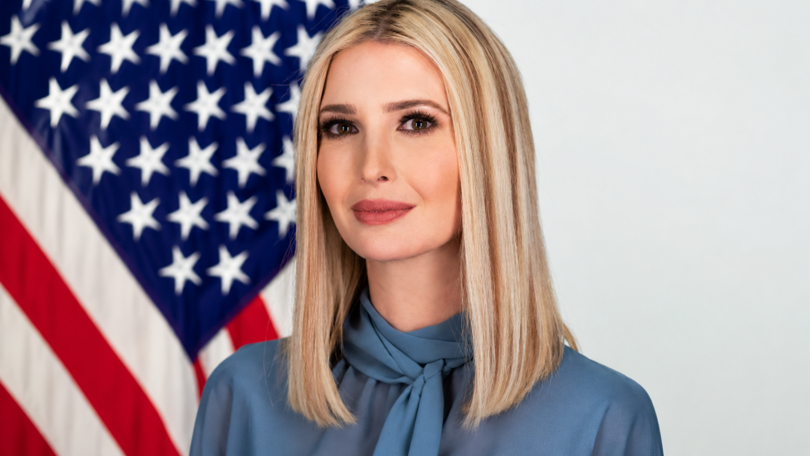 Portrait of a woman with long blonde hair wearing a blue blouse, standing in front of an American flag backdrop.