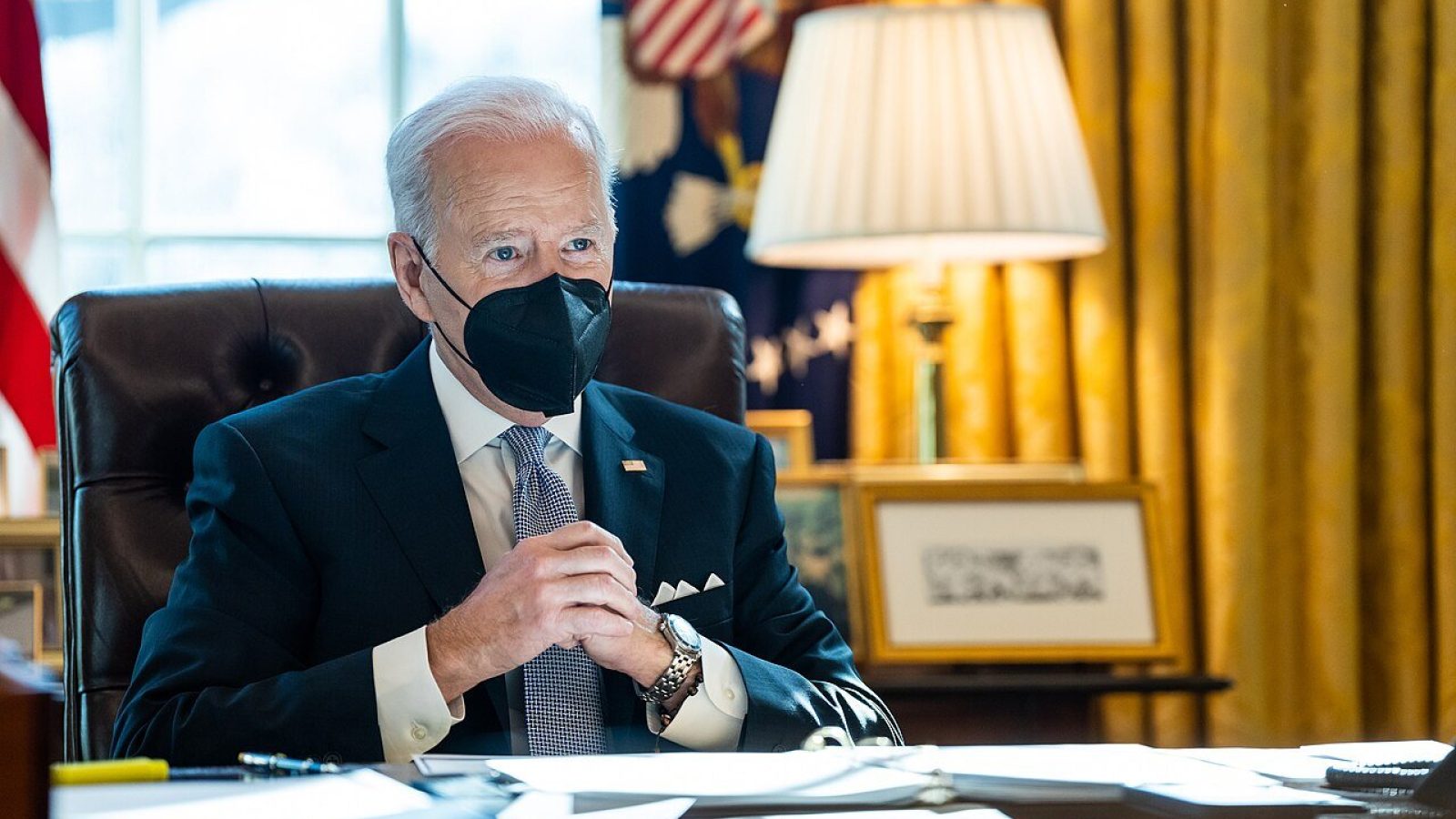 President Biden wearing a black mask sits at his desk in the Oval Office, engaged in a serious discussion with documents and a lamp visible in the background.