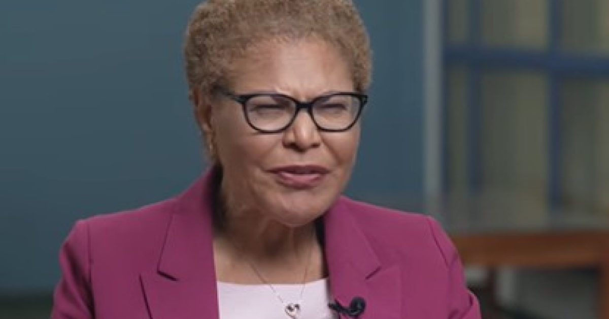 Woman with short curly hair wearing glasses and a pink blazer, speaking in a professional setting.