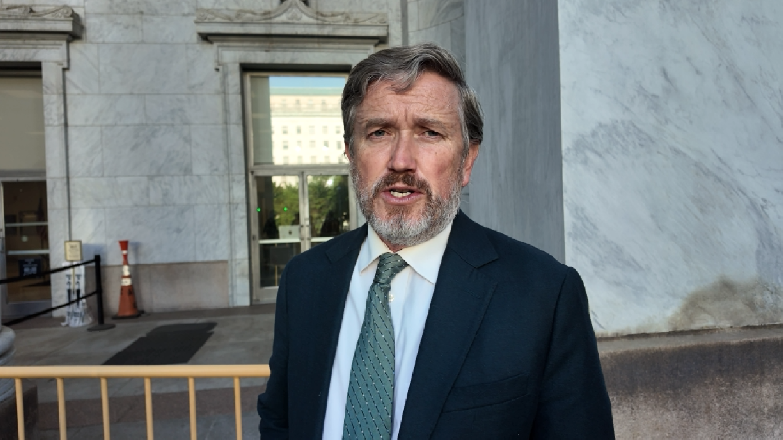 Man in a suit with a beard speaking outside a marble building, highlighting a professional setting and public engagement.