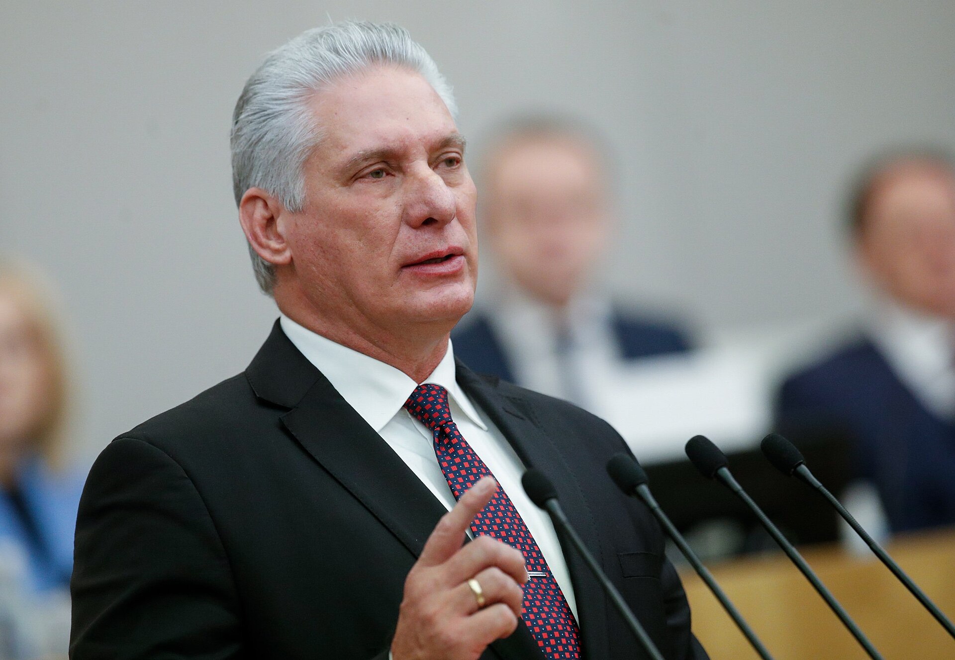 Cuban leader delivering a speech at a conference, emphasizing key points with a raised finger, dressed in a formal suit.