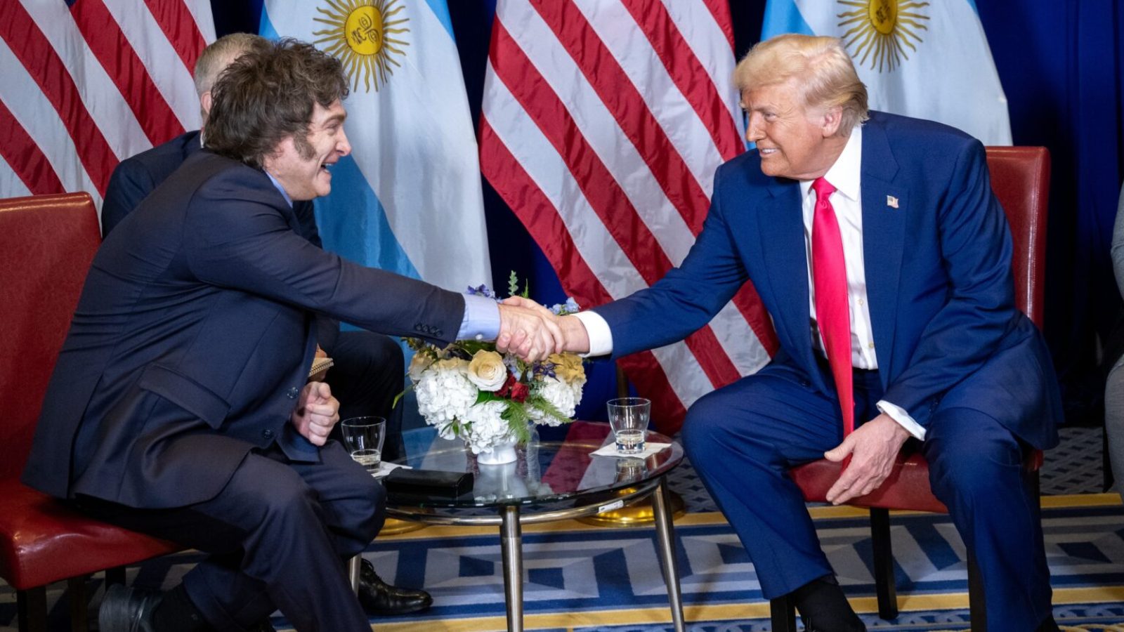 Donald Trump shakes hands with an official in front of U.S. and Argentine flags during a diplomatic meeting.