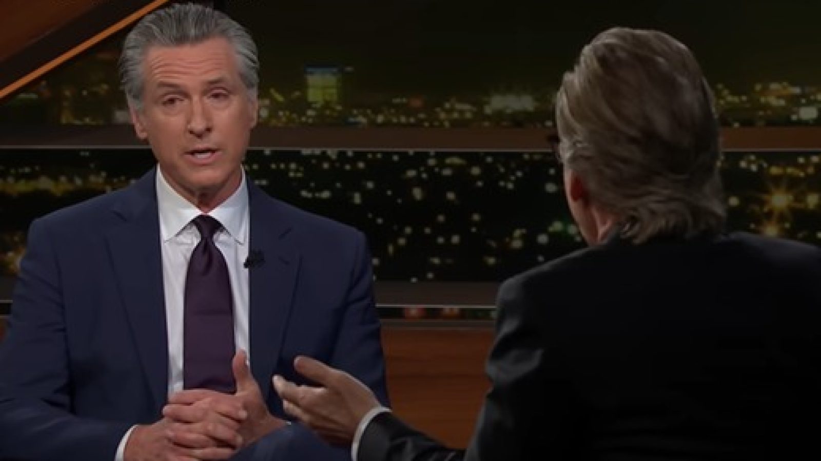 California Governor Gavin Newsom discusses key issues during an interview on a talk show, with a city skyline visible in the background.