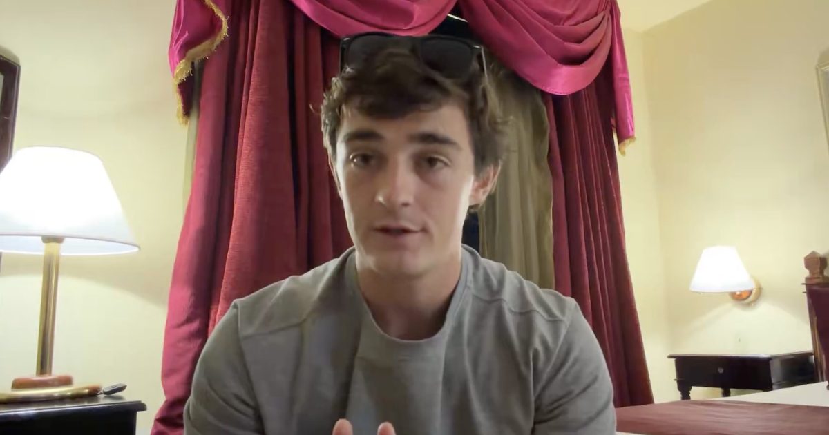 Young man in a gray shirt sitting in a hotel room with red curtains and a lamp, appearing engaged in conversation or discussion.