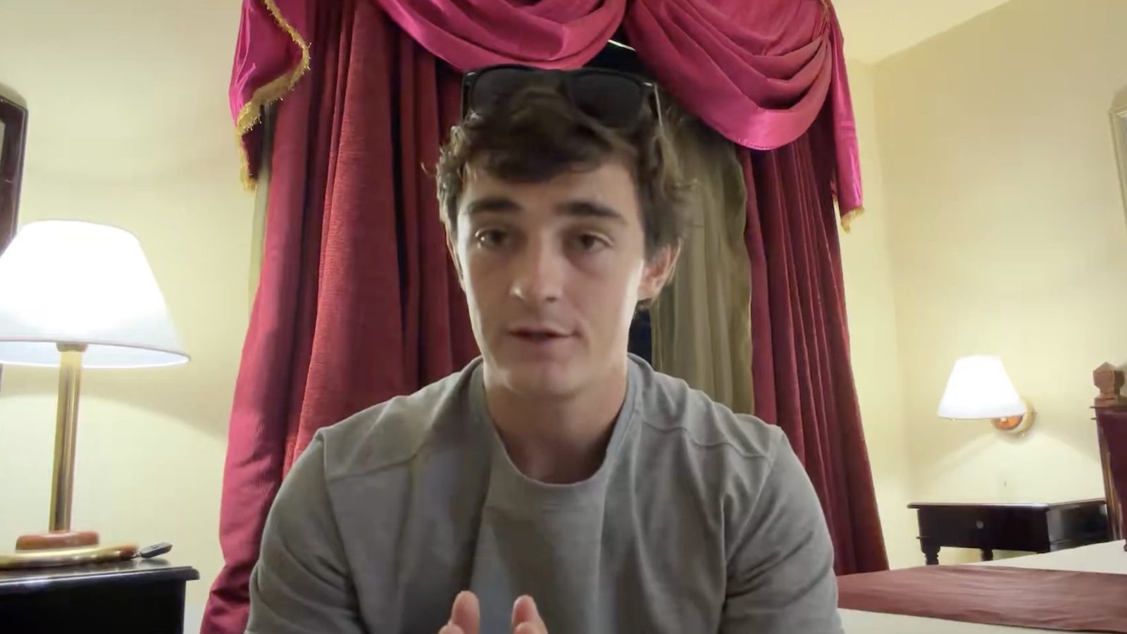 Young man in a gray shirt sitting in a hotel room with red curtains and a lamp, appearing engaged in conversation or discussion.