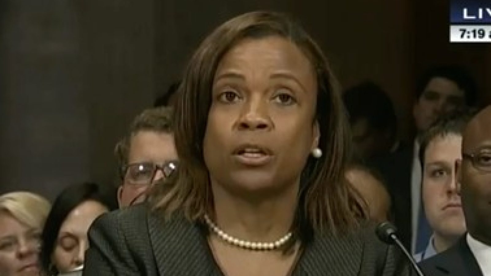A woman with brown hair and a pearl necklace speaks during a congressional hearing, surrounded by an audience of attentive listeners.