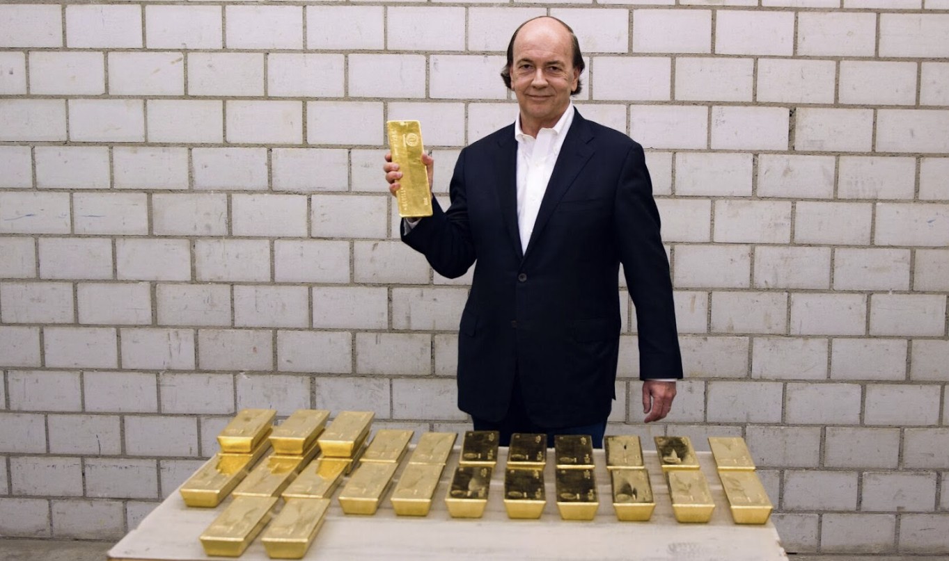 Man in a suit holding a gold bar stands in front of a table stacked with gold ingots against a brick wall backdrop.
