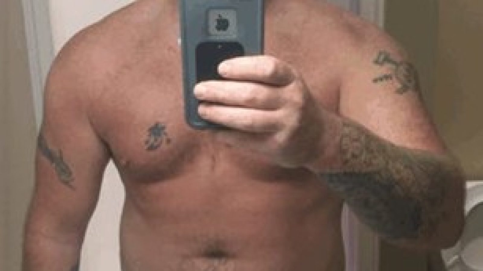 A shirtless man with tattoos takes a mirror selfie, showcasing his upper body and casual setting.