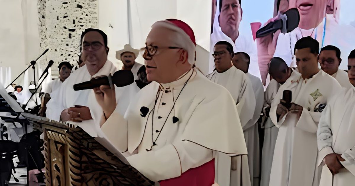 Cardinal delivers a speech during a religious ceremony, surrounded by clergy members and a large screen displaying his image.