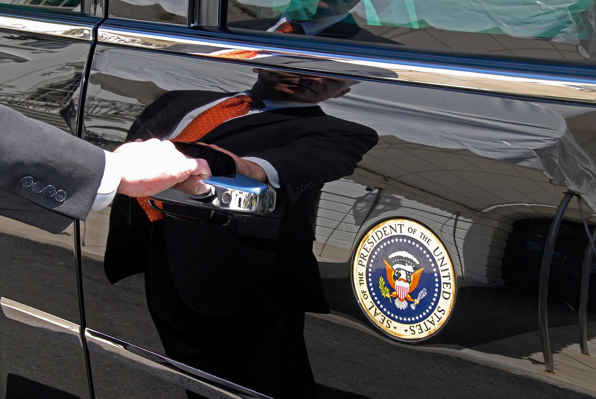 A hand reaches for the door handle of a black vehicle displaying the Presidential Seal, symbolizing official government transportation.