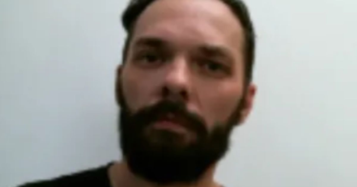 Close-up portrait of a man with a beard, wearing a black shirt, against a plain white background.