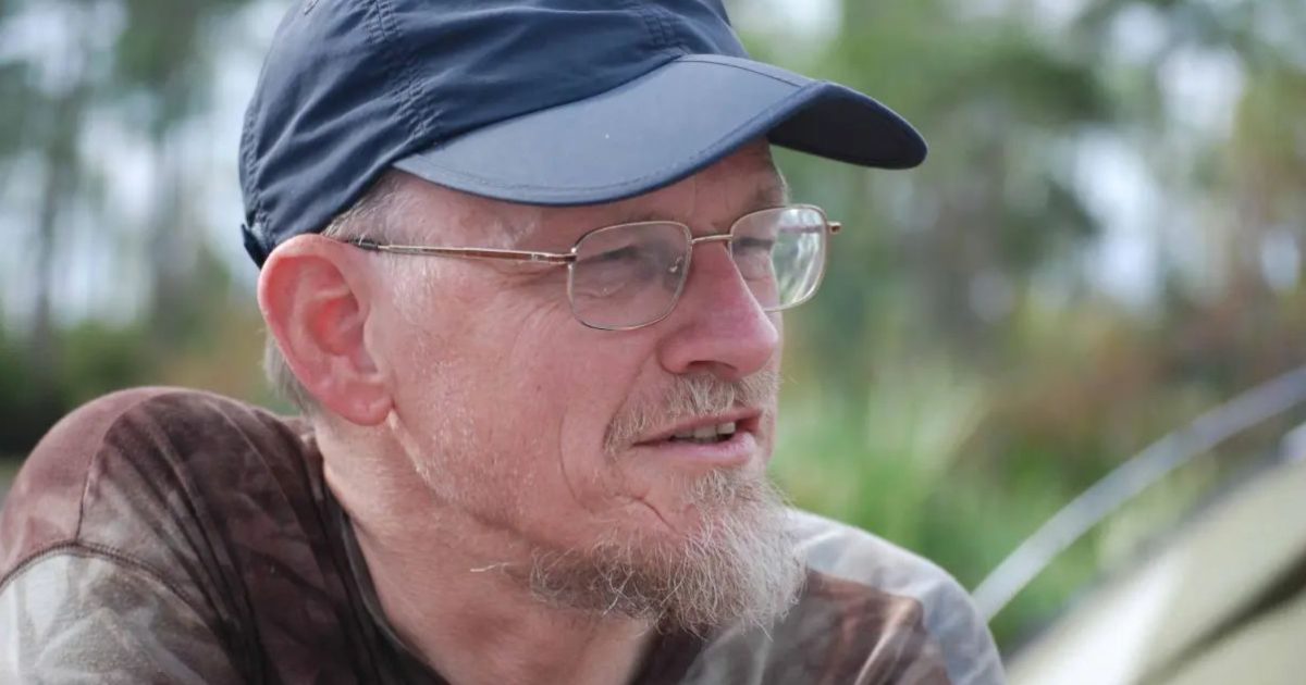 A thoughtful older man wearing a baseball cap and glasses, sitting outdoors surrounded by greenery.