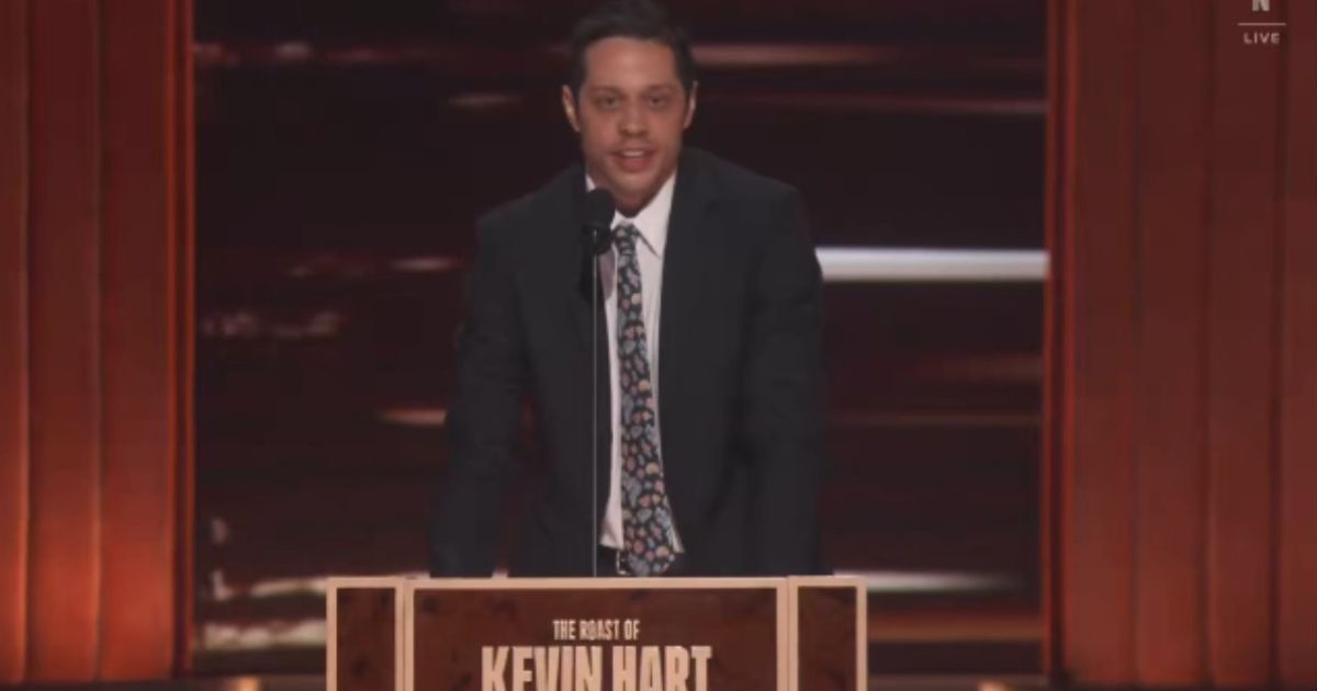 Comedian performing at the Roast of Kevin Hart, delivering jokes on stage with a microphone and a wooden podium in a well-lit setting.