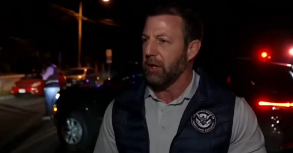 Official speaking during a nighttime press event, surrounded by emergency vehicles and police activity.