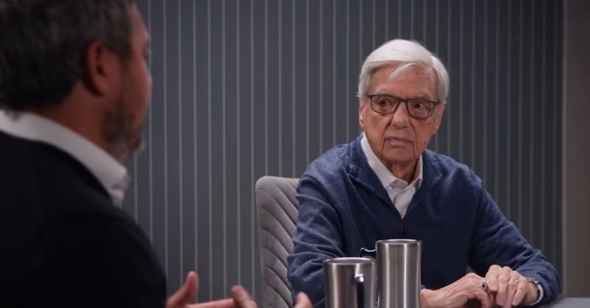 Two men engage in a thoughtful discussion at a table, with one man in a blue sweater and glasses attentively listening while the other speaks.