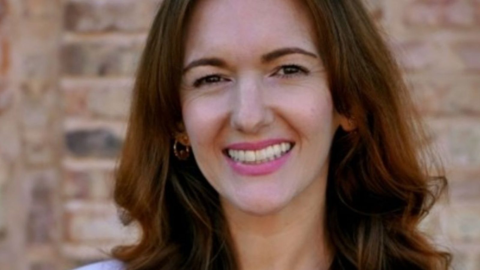 Smiling woman with long brown hair and pink lipstick poses against a brick wall, showcasing a friendly and approachable demeanor.