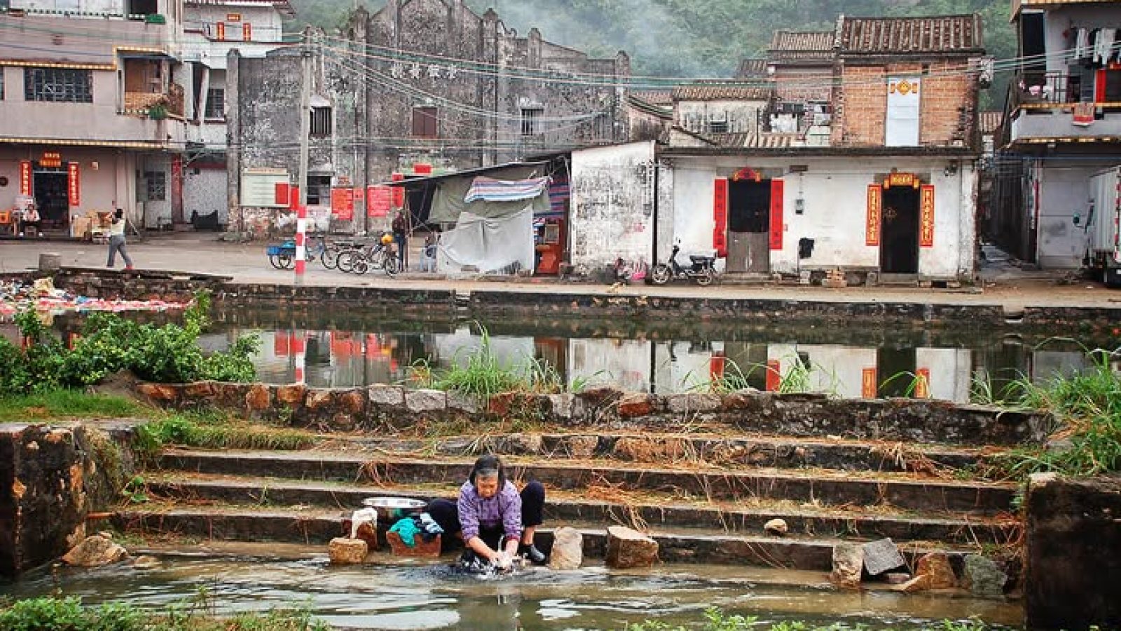 Woman washing clothes by a tranquil waterway in a traditional village setting, surrounded by rustic buildings and lush green scenery.