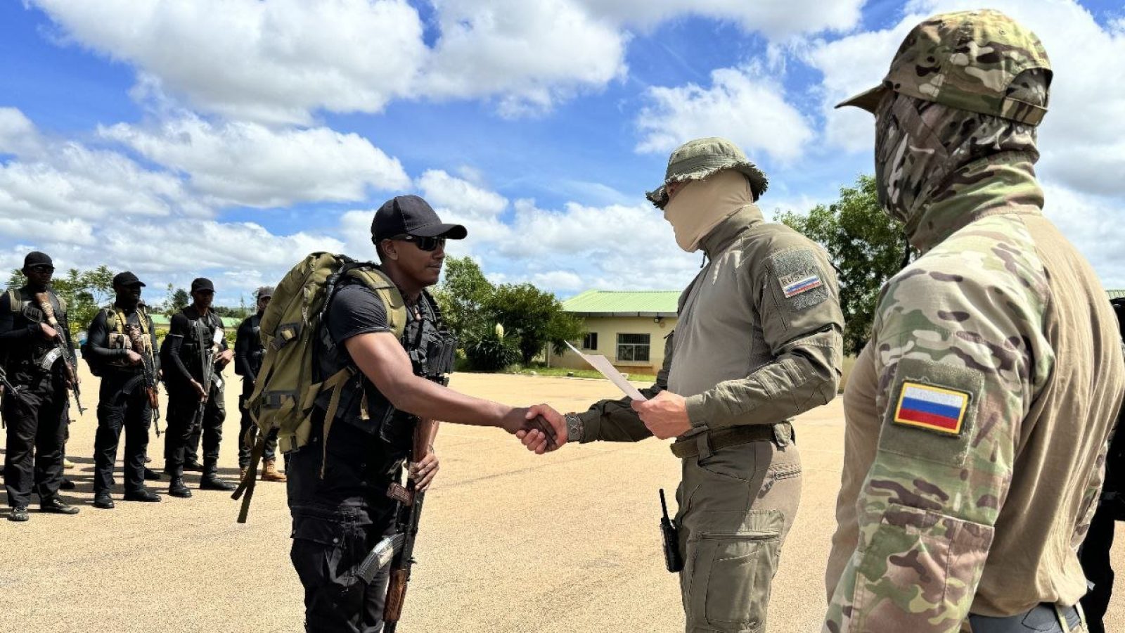 Military personnel engage in a formal handshake during a training exercise, with armed soldiers in the background, highlighting teamwork and collaboration.
