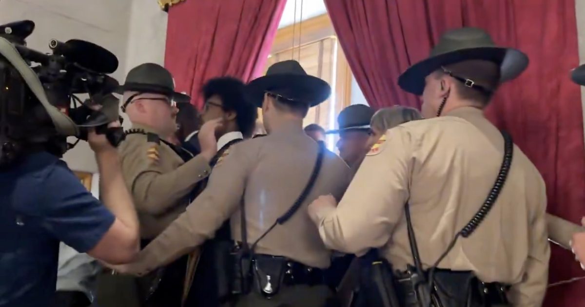 Law enforcement officers engage in a tense confrontation with individuals in a formal setting, with cameras capturing the scene.
