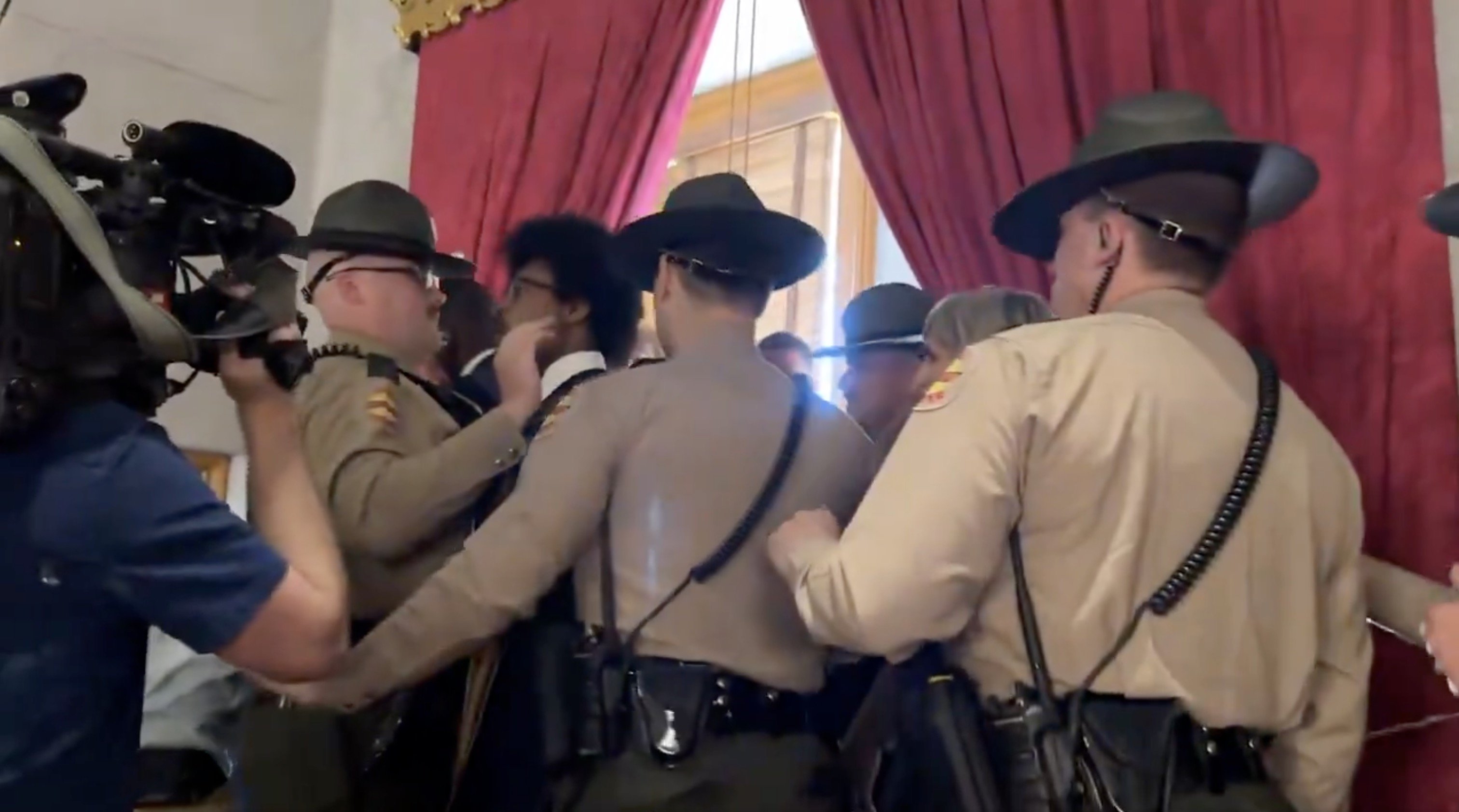 Law enforcement officers engage in a tense confrontation with individuals in a formal setting, with cameras capturing the scene.
