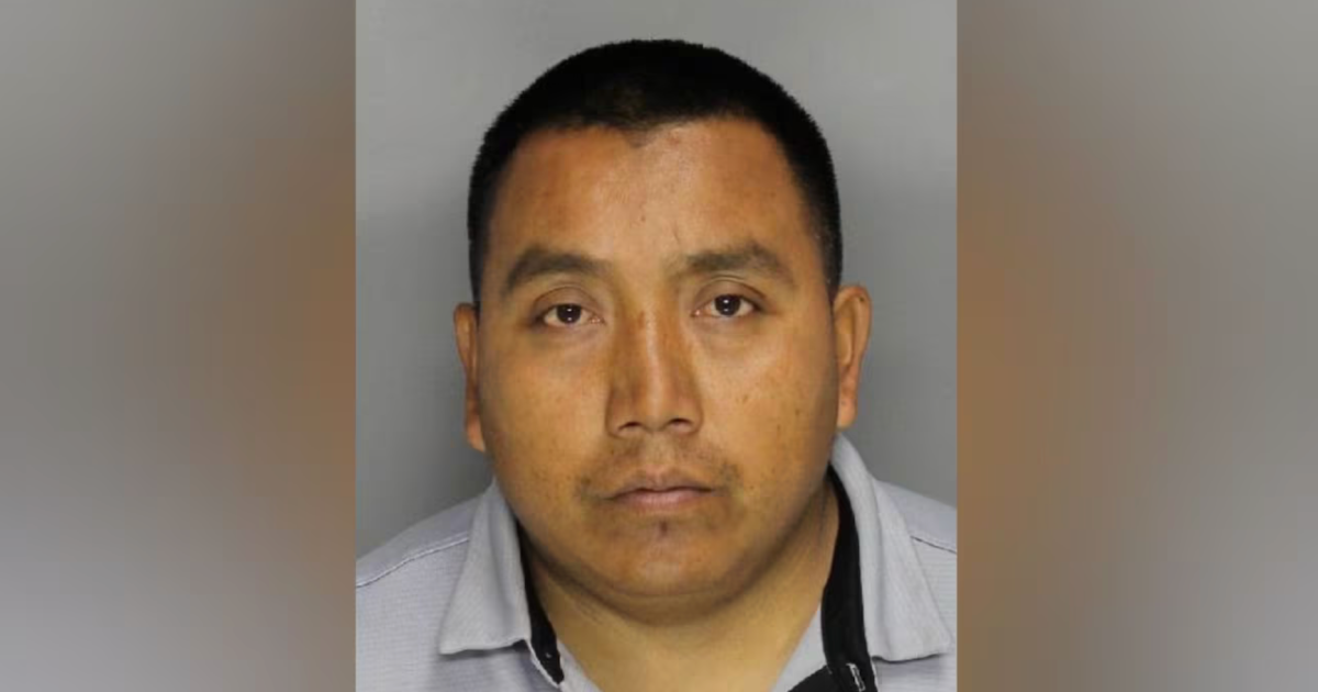 Mugshot of a male suspect with short dark hair wearing a gray collared shirt, staring directly at the camera against a neutral background.