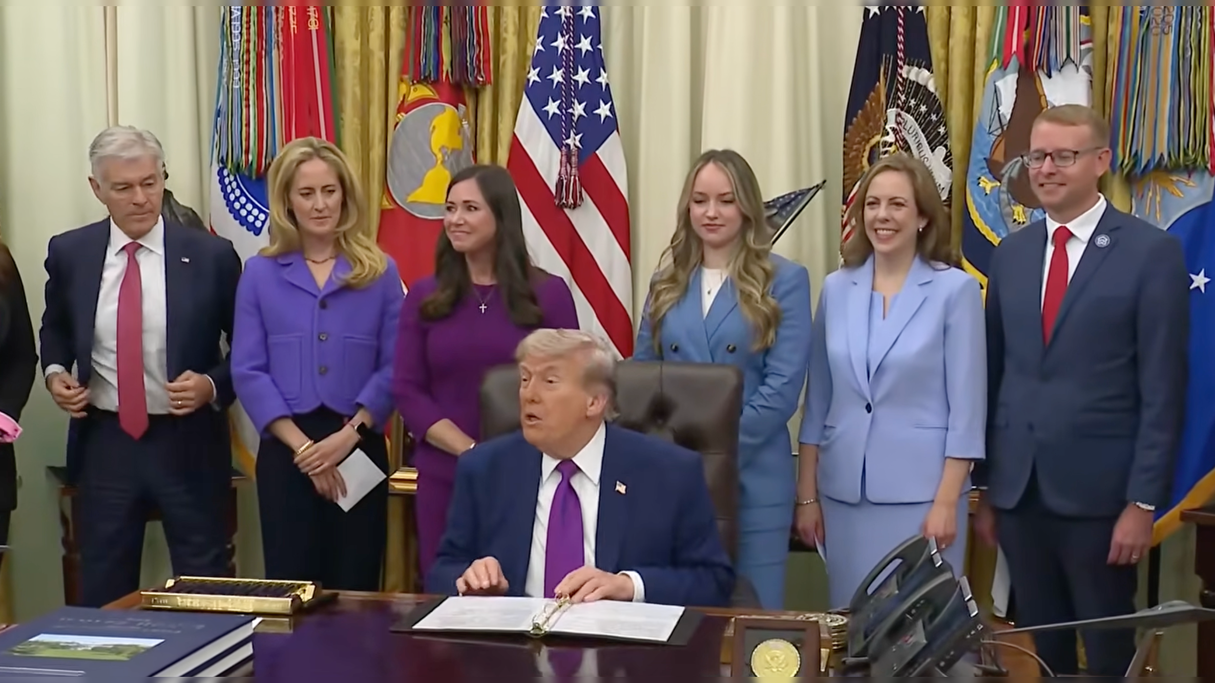 Donald Trump seated at a desk in the Oval Office, surrounded by six individuals, with U.S. flags and military insignia in the background.