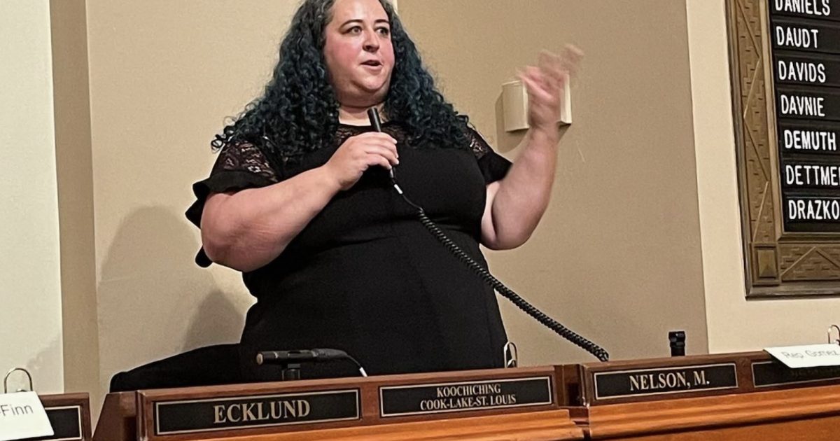 A woman with curly hair speaks passionately at a podium during a meeting, emphasizing key points with hand gestures.