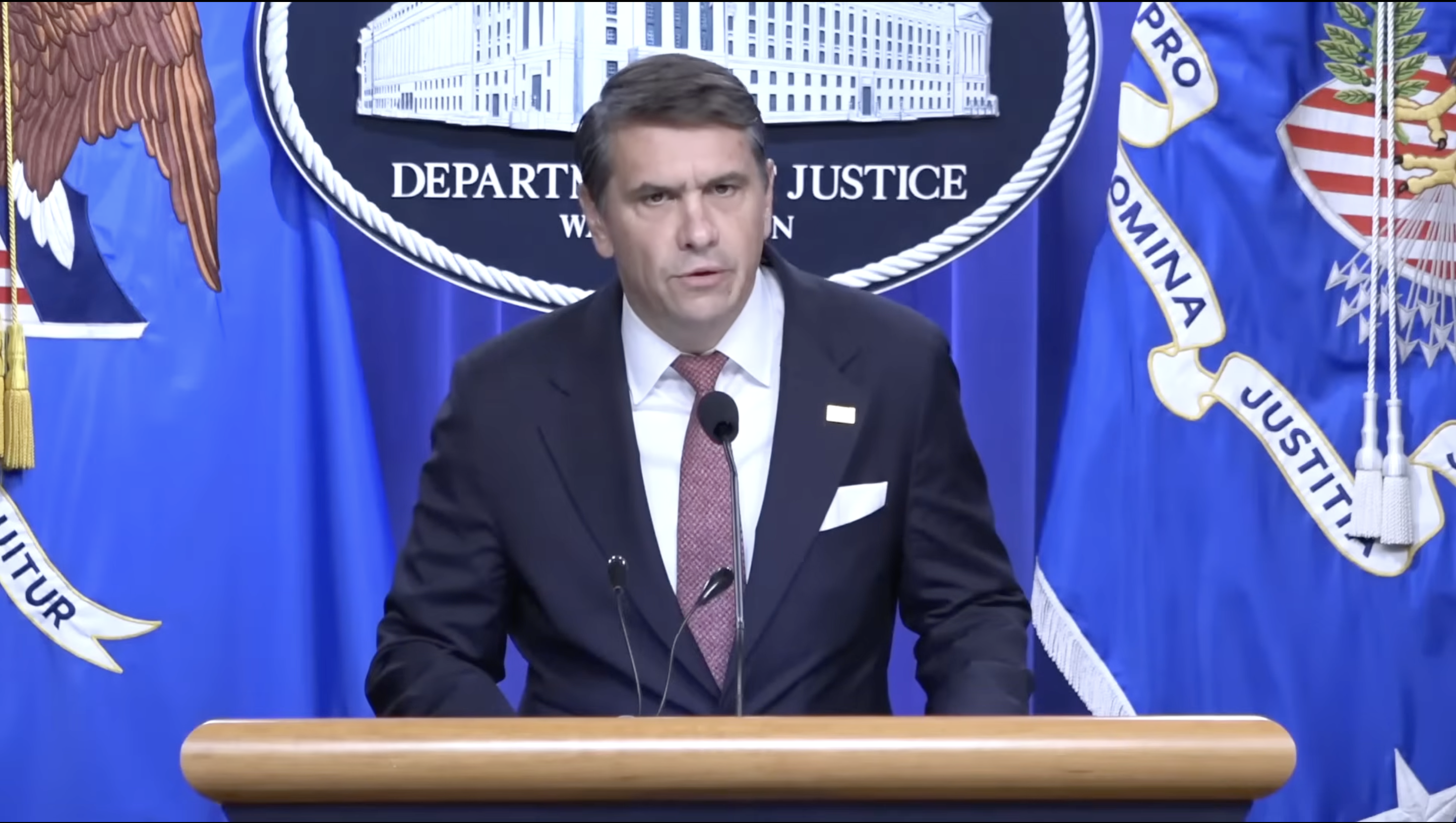 Official speaking at a Department of Justice press conference, with flags and emblems in the background, addressing important legal matters.