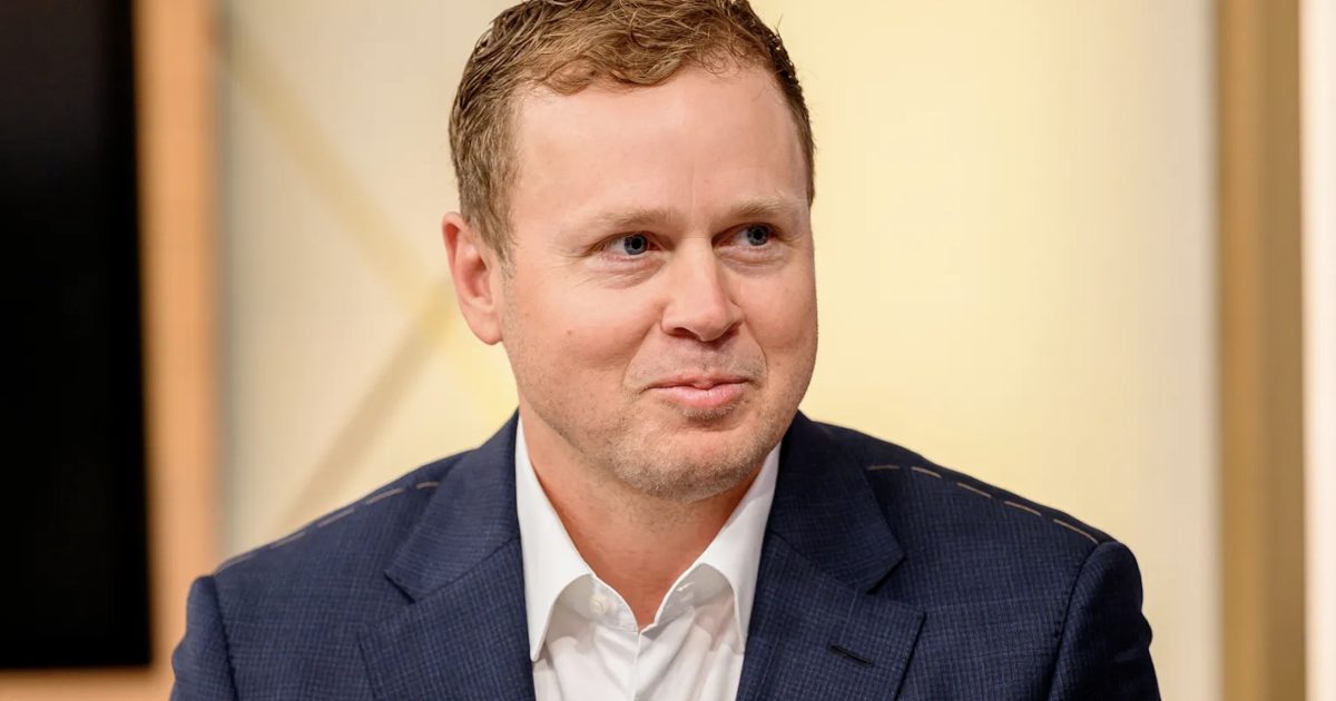 A smiling man in a blue suit and white shirt sits against a soft-focus background, exuding confidence and approachability.