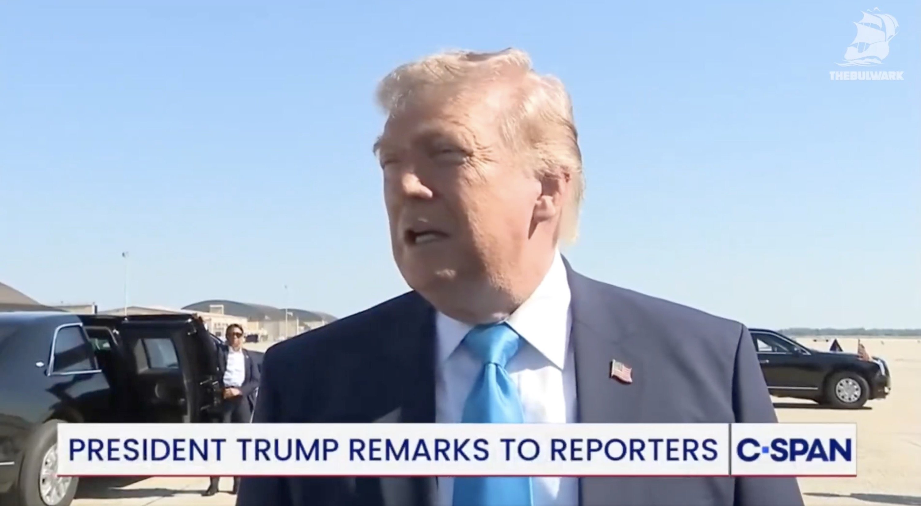 Donald Trump speaking to reporters outside near a black vehicle, with clear blue skies in the background.