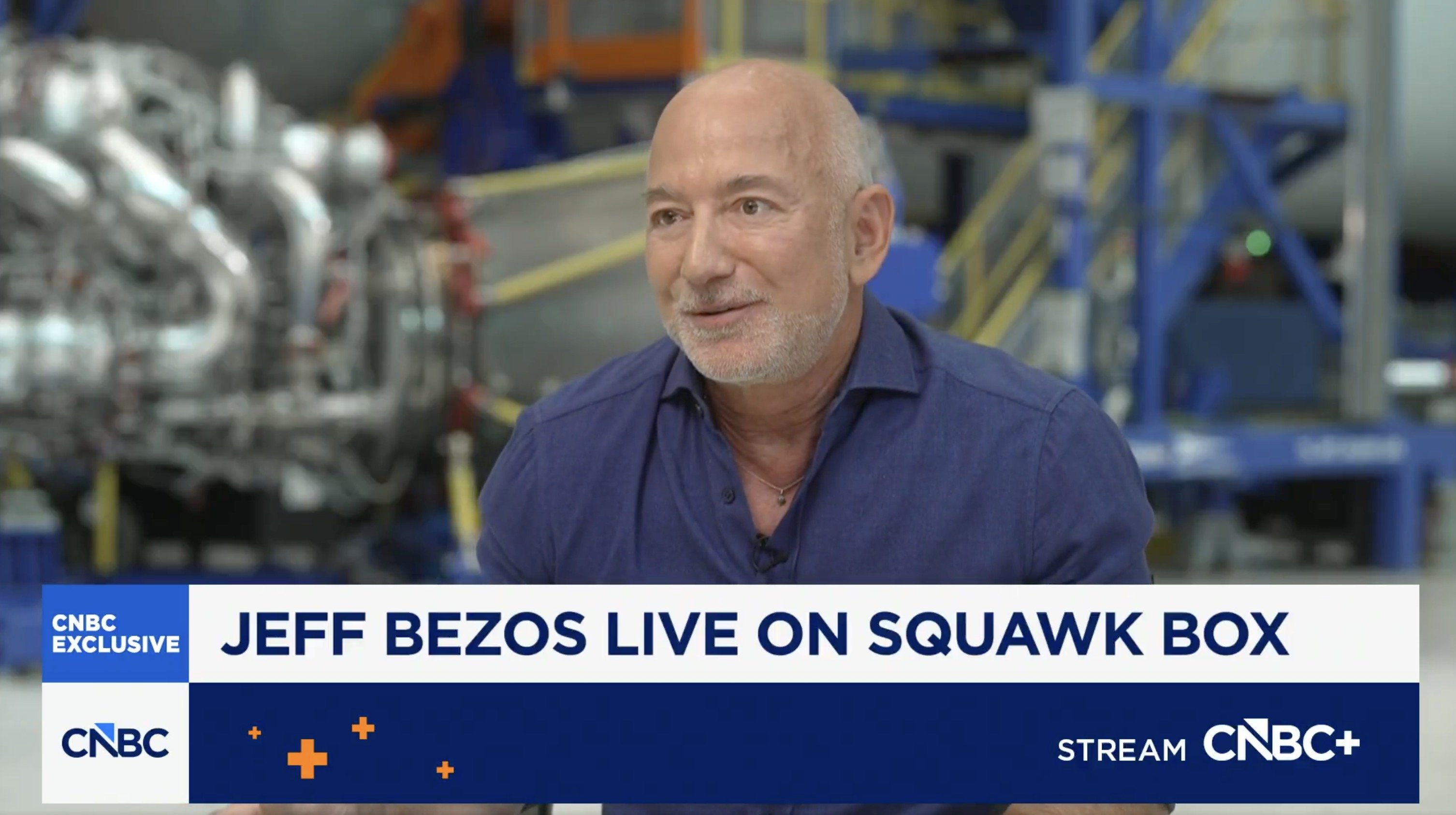 Jeff Bezos speaking during a CNBC interview in a high-tech facility, featuring aerospace equipment in the background.