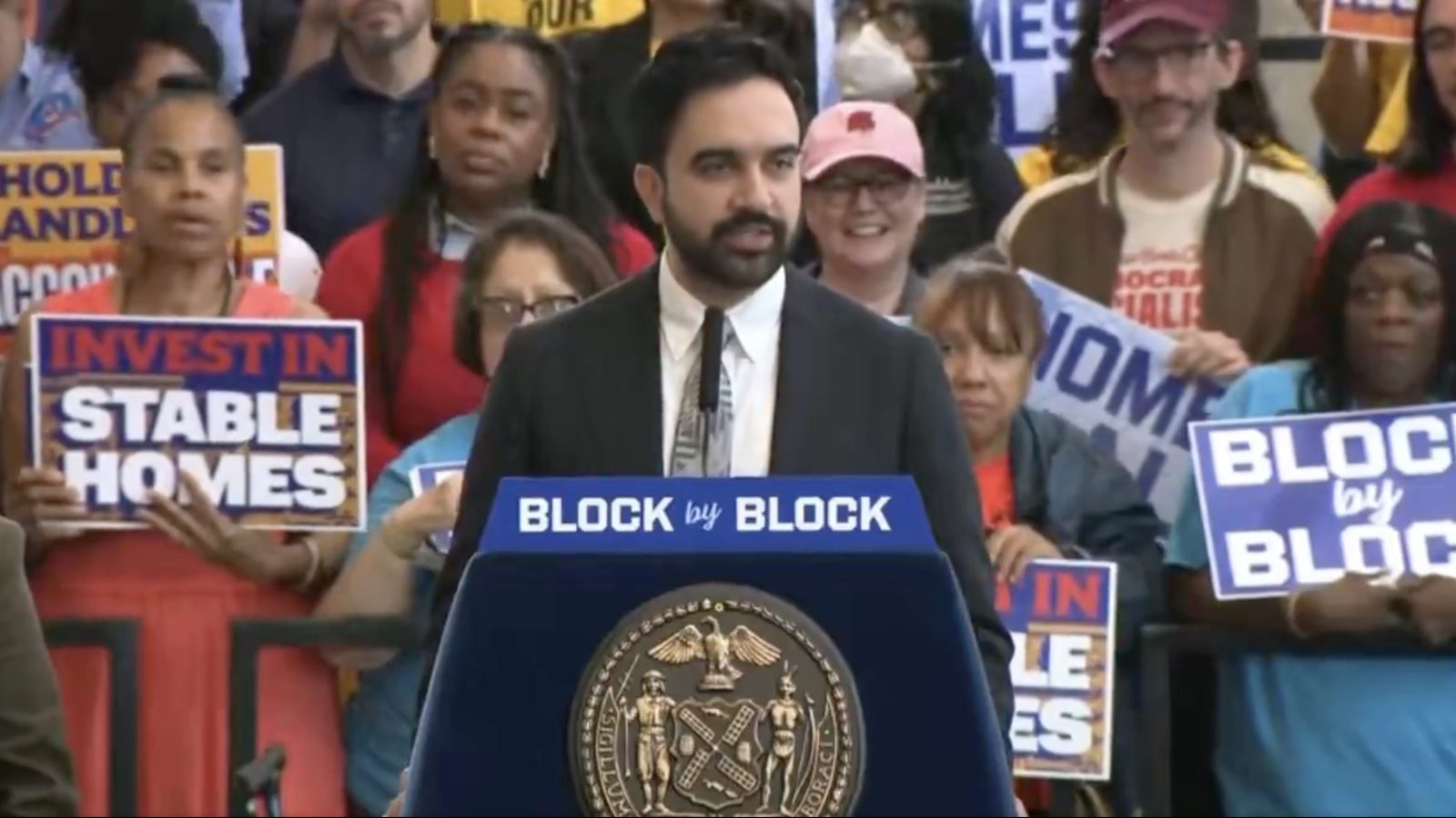 A speaker addresses a crowd advocating for stable homes and community investment during a public event, with attendees holding supportive signs.