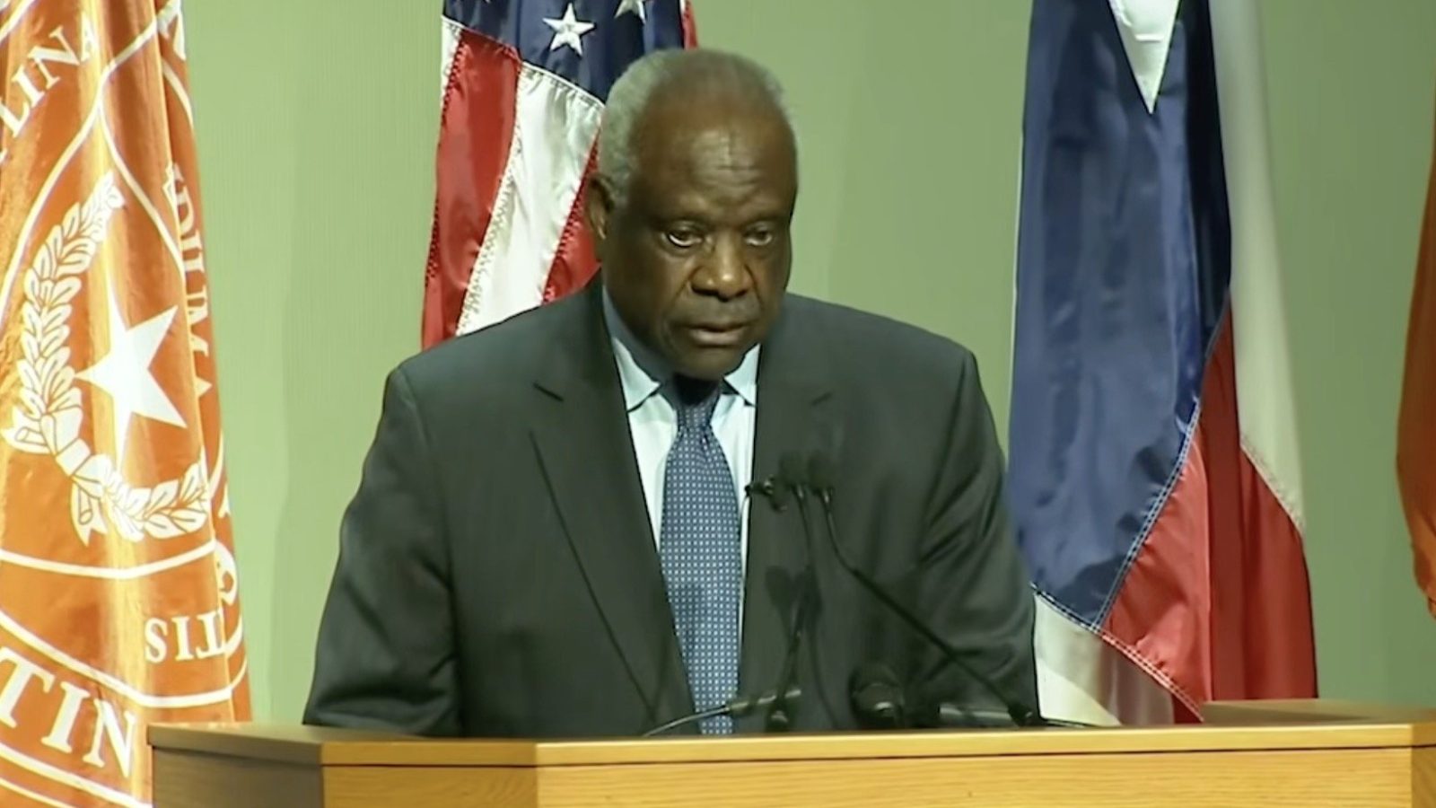 A man in a suit speaks at a podium with flags in the background during a formal event.