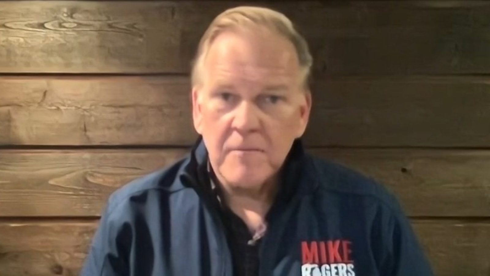 Mike Rogers speaking in front of a wooden backdrop, wearing a blue jacket with his name and campaign logo prominently displayed.
