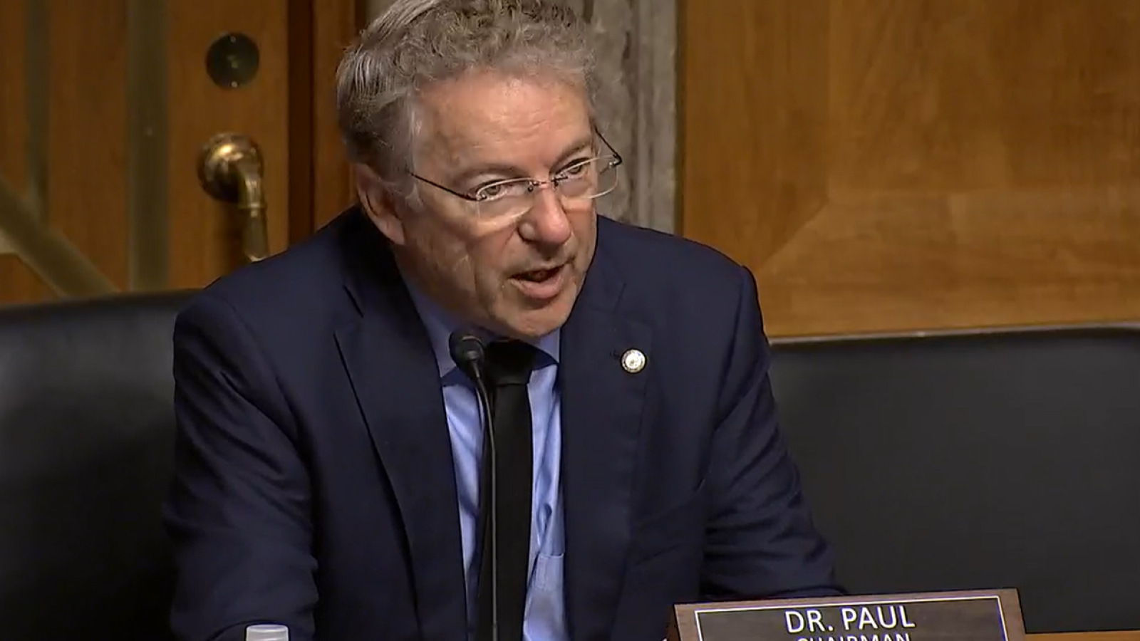 Dr. Paul, the Chairman, speaks during a Senate committee meeting, emphasizing key issues while seated at a desk with a nameplate.